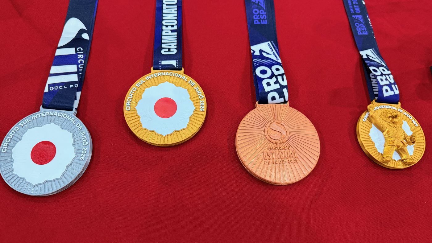 Quatro medalhas do Campeonato Estadual de Judô 2026 em prata, ouro, cobre e a versão kids, com fitas azuis, sobre toalha vermelha.