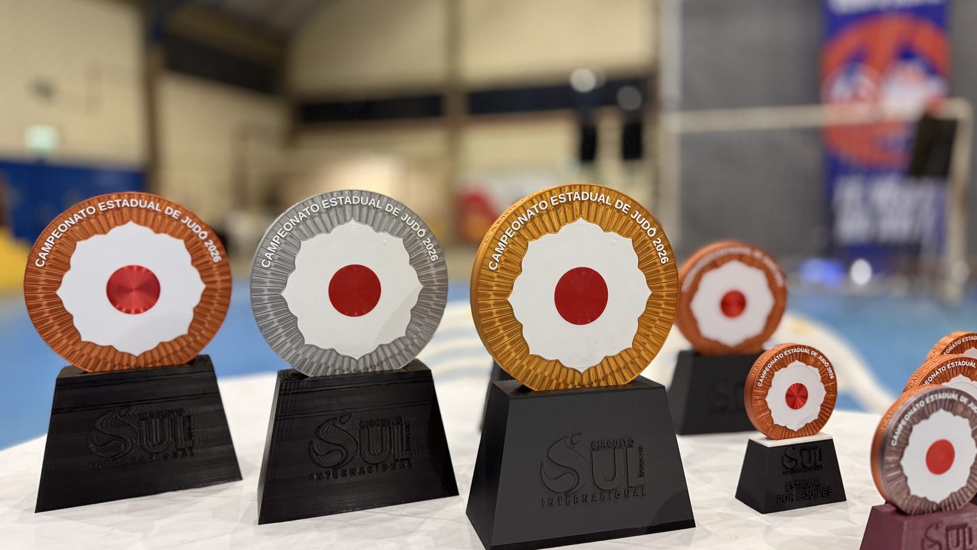 Três troféus de disco em cobre, prata e ouro com hinomaru em relevo, sobre bases pretas no ginásio do Campeonato Estadual de Judô 2026.