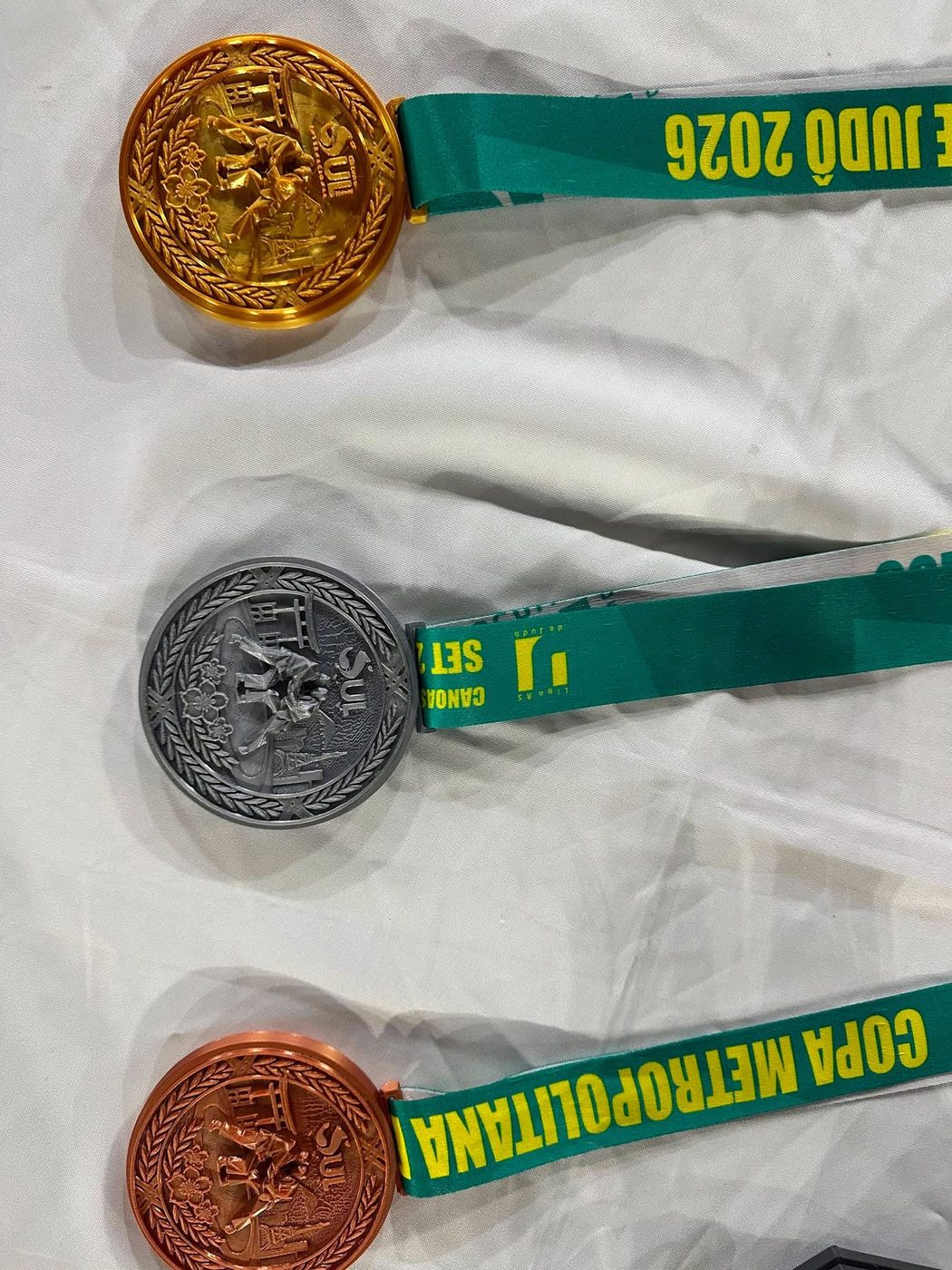 Medalhas de ouro, prata e cobre com fitas verdes da Copa Metropolitana 2026, sobre tecido branco.