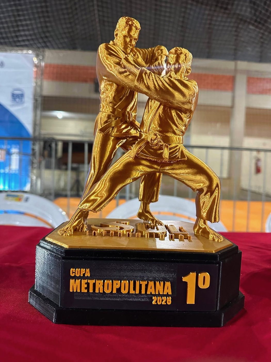 Troféu Tai Otoshi dourado com a placa de 1º lugar da Copa Metropolitana 2026, sobre a mesa de premiação no ginásio.