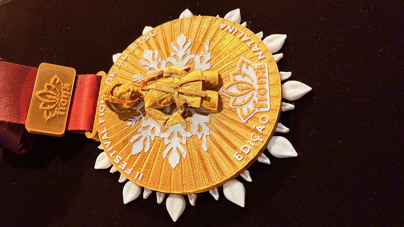 Medalha do Festival Fiona de Jiu-Jitsu deitada, dourada com flocos brancos, passador com a marca Fiona e fita vermelha.