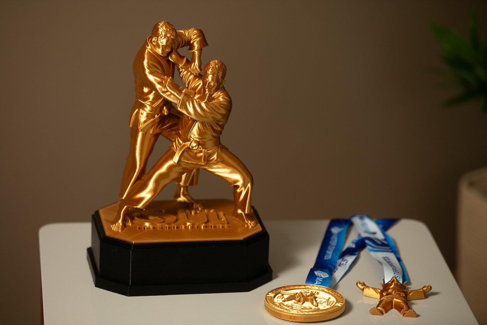 Troféu Tai Otoshi dourado ao lado de uma medalha e de uma medalha kids com mascote, sobre mesa branca.
