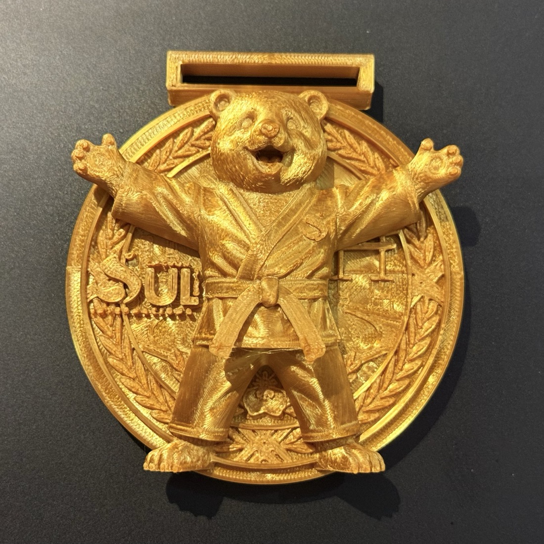 Medalha kids dourada vista de frente, com o mascote urso judoca sobre a coroa de louros.