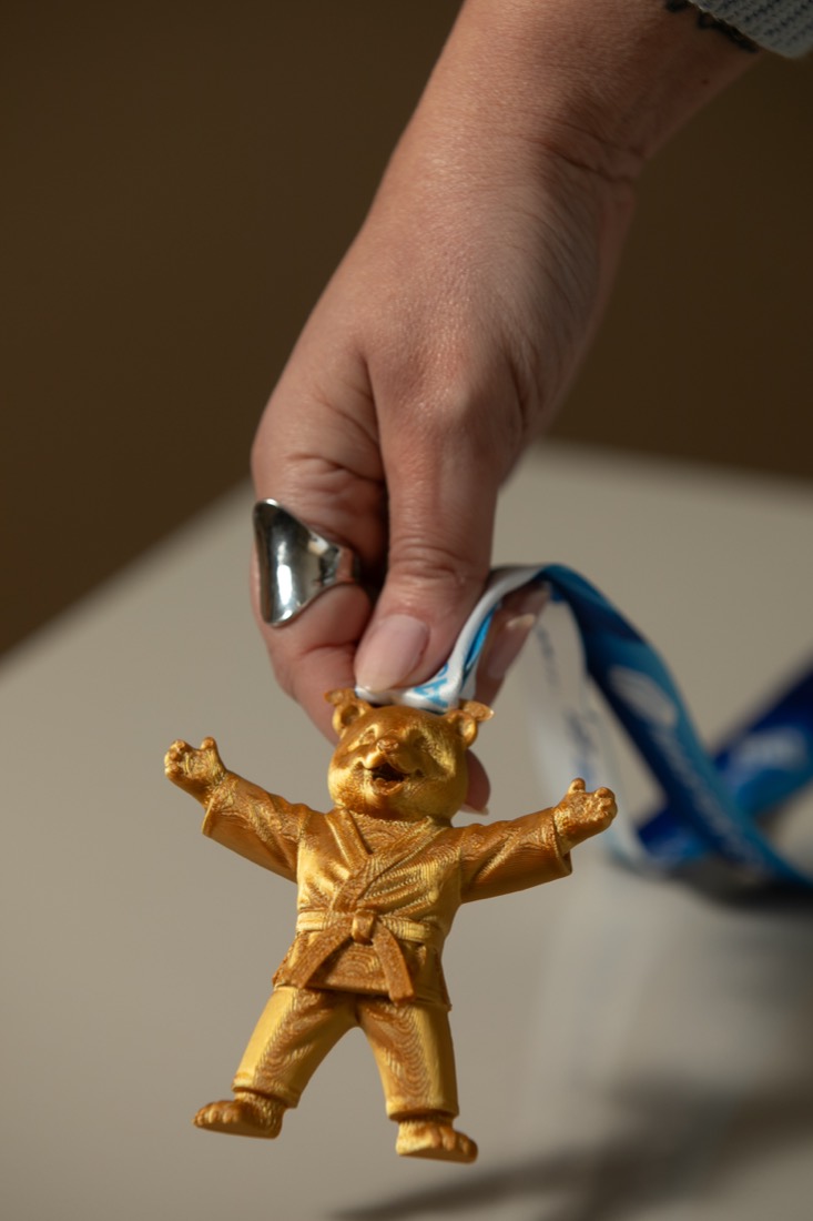 Mão segurando pela fita a medalha kids com o mascote urso judoca de braços abertos.