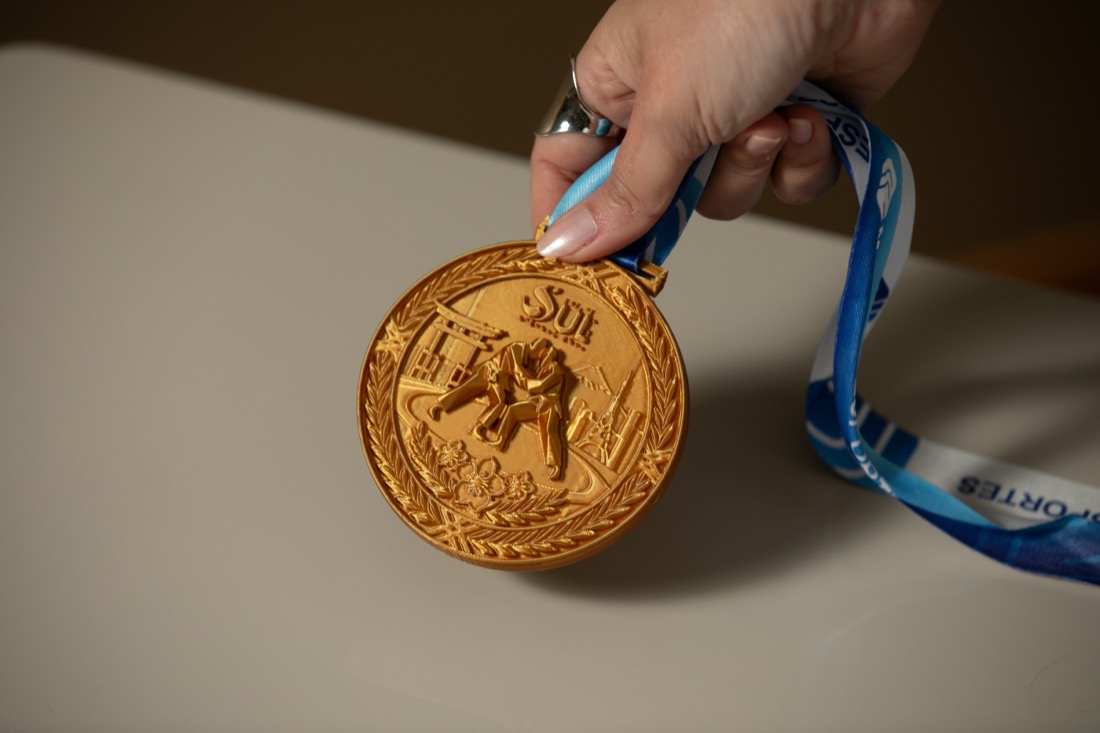 Mão segurando uma medalha 3D dourada pela fita, mostrando a escala da peça.