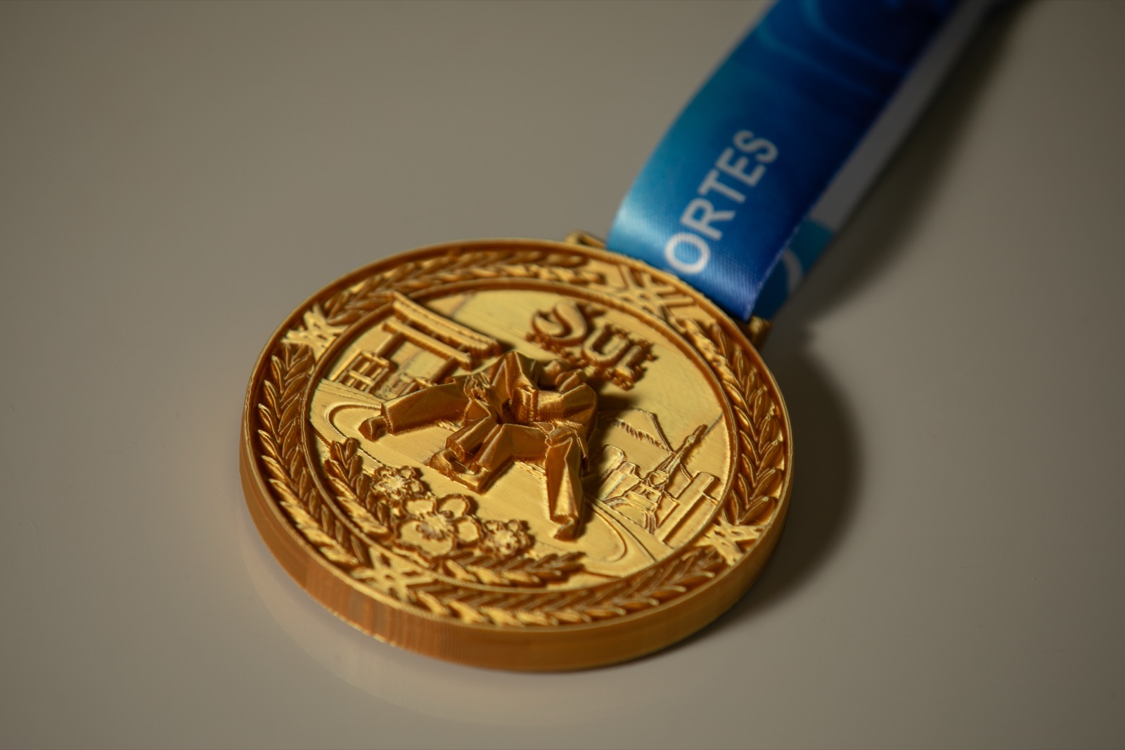 Medalha 3D dourada com dois judocas em alto relevo, torii e coroa de louros, com fita azul da Master Esportes.