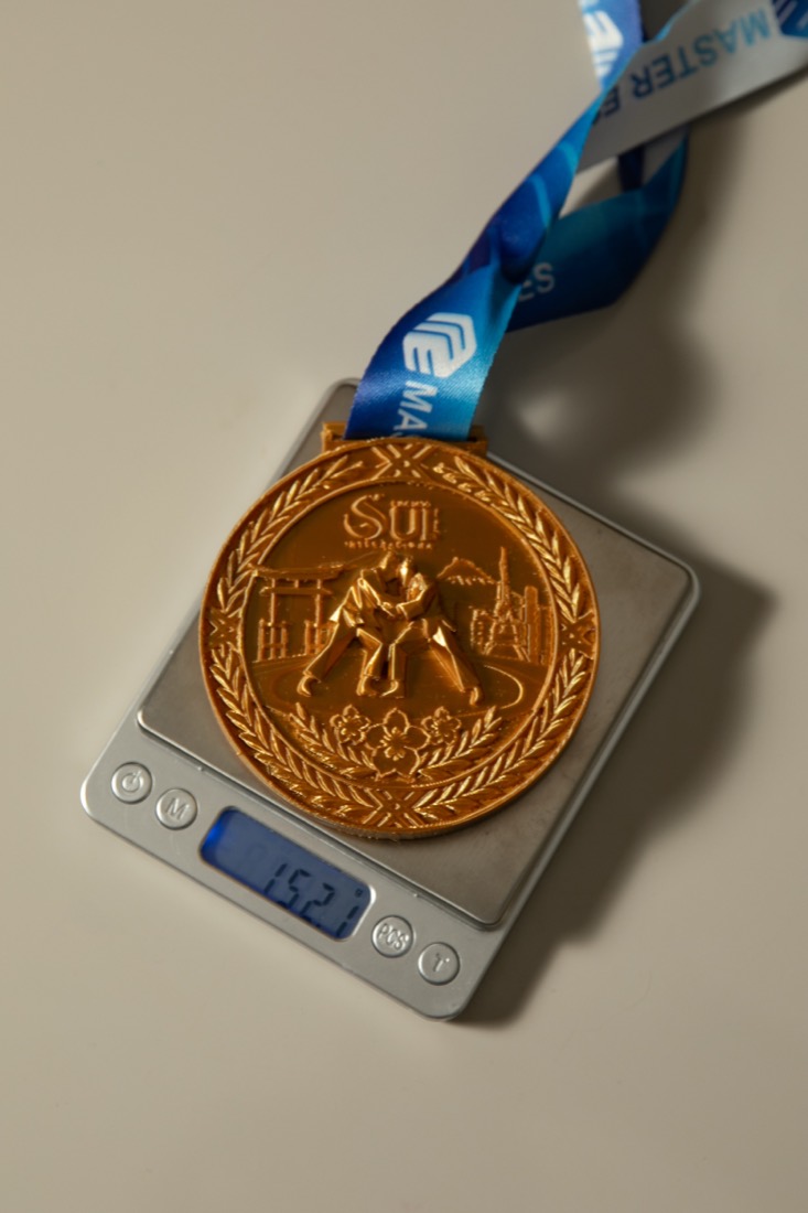 Medalha dourada sobre balança digital marcando 152 gramas.
