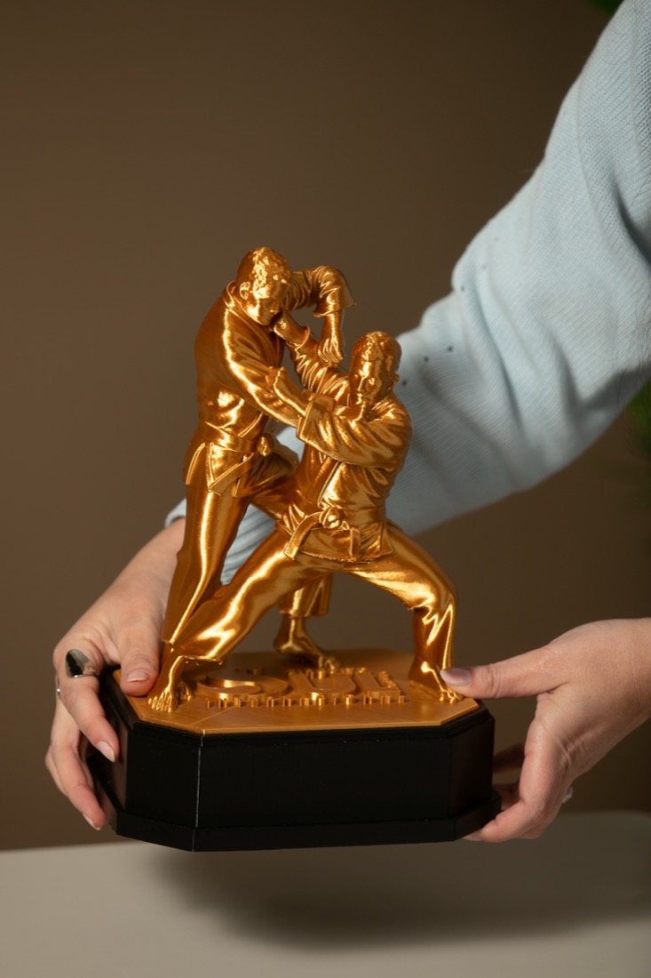 Duas mãos segurando o troféu Tai Otoshi pela base, mostrando o tamanho real da peça.