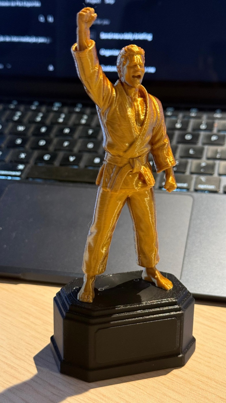 Troféu Vitória Full dourado: judoca de judogi com o punho erguido em comemoração, sobre base octogonal preta.