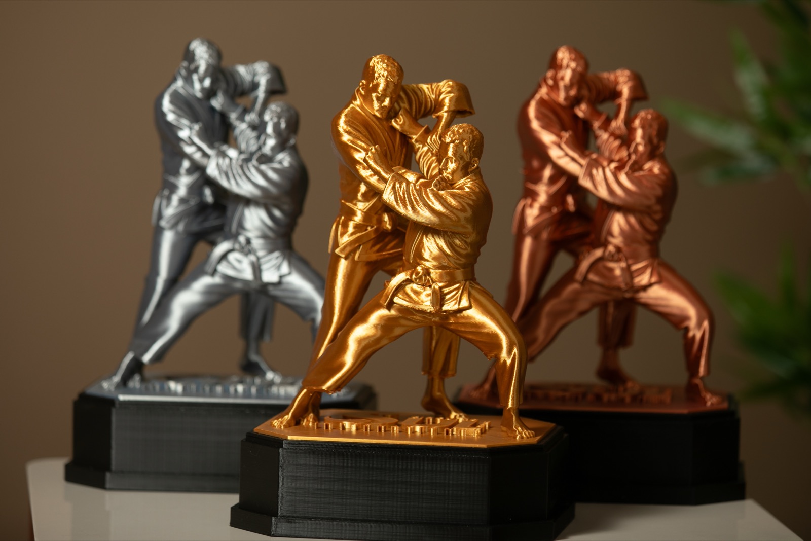 Três troféus Tai Otoshi lado a lado nos acabamentos ouro, prata e cobre, sobre bases pretas.